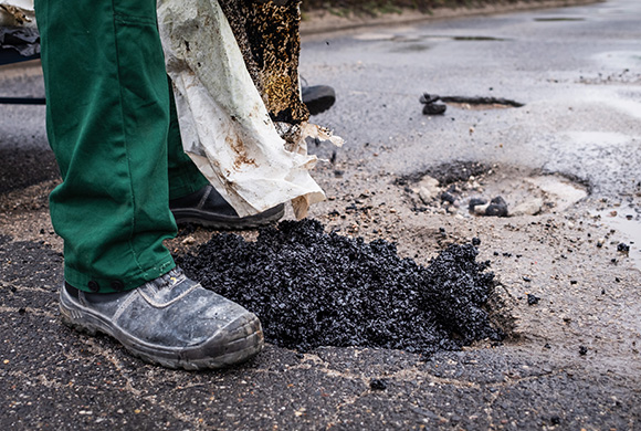 worker repairing pothole with asphalt mixture on road surface near foot wearing safety boot six repairs needed