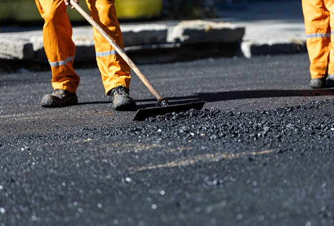 person in orange workwear using a shovel to spread asphalt on a road construction site with focused attention on asphalt application process